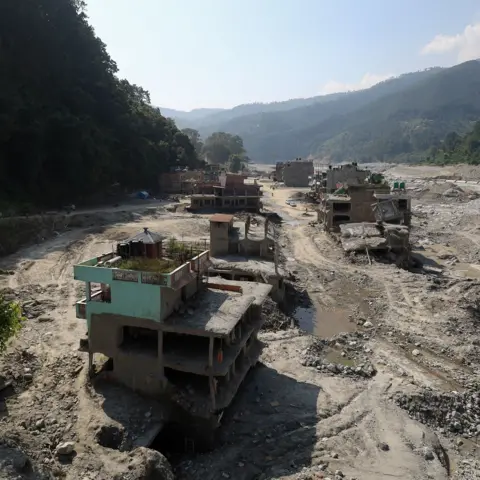 Subaas Shrestha/NurPhoto via Getty Images Flood-damaged buildings in a vast landscape of mud, gravel and debris in Trishuli Bazar, Nepal, nearly two months after devastating floods swept through the valley. Several structures are partially collapsed or left stranded above eroded ground, while a widened river channel and scarred floodplain stretch through the settlement between steep, forested hills.