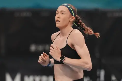 Getty Images Joanna Wietrzyk wears a black sports bra and a dark headband with the words 'the feed' printed in green letters. Her hair is tied back into a plait and she is mid-run.