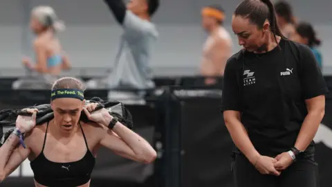 Getty Images Joanna Wietrzyk competing in Hyrox competition lifting a heavy bag as an official looks on. 