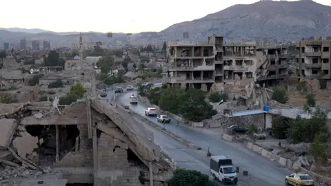 Neha Sharma/BBC Wide shot shows a built up urban area with several tall buildings seriously damaged, a number of cars driving on a road running through the centre and mountains in the background