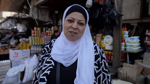Neha Sharma/BBC A woman in her 50s wears a white hijab and looks to the camera, in front of an outdoor market stand selling food items