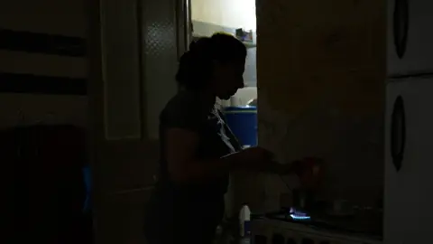 Neha Sharma/BBC Profile of a woman heating up water on a stove standing in near darkness