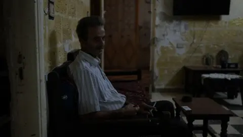 Neha Sharma/BBC A man wearing a striped shirt sits in his living room in darkness