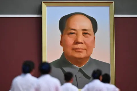 AFP via Getty Images Large framed portrait of Mao Zedong hanging on a red wall, viewed from below with several people in light-coloured clothing standing and facing the portrait in the foreground.