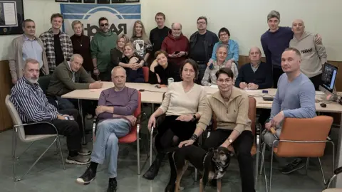 Novaya Gazeta About 20 journalists from Novaya Gazeta gathered around a table looking forward. One has a dog next to them