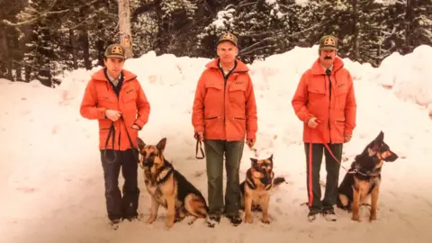 Supplied/Scott Ward An image showing three dog handlers as part of the Canine rescue team, with their dogs beside them. All three dogs are German Shepherds, and all three handlers are dressed similarily in a bright orange jacket, a park warden hat and dark trousers.