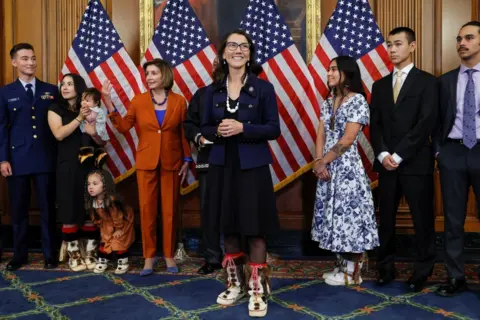 Reuters Mary Peltola poses for the cameras after her swearing-in ceremony in front of several US flags. She is wearing traditional boots of the Yup'ik people. Family members and supporters stand around her, including adults and children, some wearing traditional-style footwear and clothing. Nancy Pelosi, in an orange trouser suit, stands nearby.