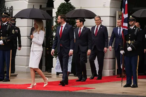 Getty Images Members of the US cabinet arrive ahead of the ceremonial welcome for King Charles and Queen Camilla on the South Lawn of the White House. A woman in a white skirt suit carries a black umbrella, while (left to right) Vice-President JD Vance, Secretary of State Marco Rubio, Secretary of the Treasury Scott Bessent and Secretary of War Pete Hegseth follow. All are dressed in dark suits with red ties, except for Hegseth who is wearing a striped blue tie. 