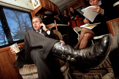 Getty Images Former US president George W Bush seated inside a campaign-style bus, wearing a dark suit and patterned tie while talking on a mobile phone and holding a paper coffee cup. One leg is stretched forward, showing a black cowboy boot decorated with a Texas flag motif. Other people are visible in the background reviewing documents.