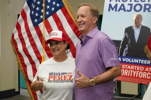 Facebook/KenPaxtonTX Ken Paxton and a female supporter pose in front of a large US flag and a campaign poster featuring Donald Trump. The woman wears a white campaign T-shirt and red-and-white baseball cap. Paxton wears a purple polo shirt. Both give a thumbs-up gesture.