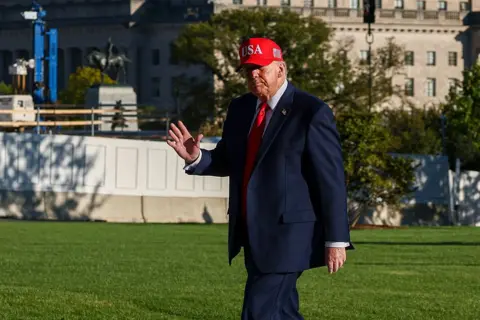 Getty Images US President Donald Trump walks across a lawn wearing a navy suit, white shirt, red tie and a red cap with "USA" embroidered on the front. One hand is raised in a wave.