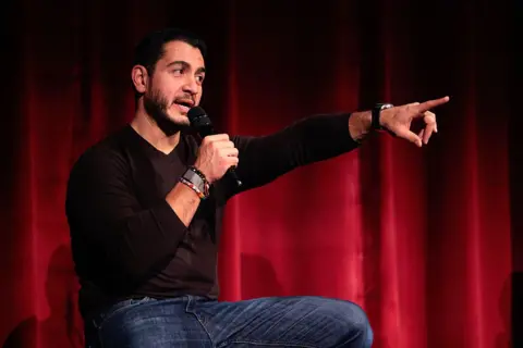 Getty Images Abdul El-Sayed sits on a stage in front of a red curtain, wearing a black V-neck, long-sleeved T-shirt and blue jeans. Holding a microphone in one hand, he gestures with the other while speaking.