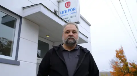 BBC/Nadine Yousif An image showing a man, Stephane Lefebvre, outside a small building with a sign that reads 'UNIFOR Syndicat Quebec'. The man is older with grey hair and a beard, is looking right at the camera and is wearing a black zip up jacket on top of a dark grey polo shirt.