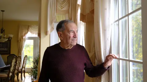 BBC/Nadine Yousif An image showing Frank Dottori inside his home, looking outside of a window. His face is illuminated by the natural light outside. He is older and has grey hair, and is wearing a deep burgundy sweater. Behind him are parts of his home dining room visible and curtains. 