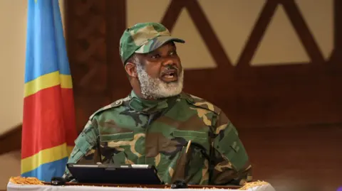 Getty A man with white beards standing on a podium wearing combat uniform and a matching cap infront of a blue, yellow and red flag