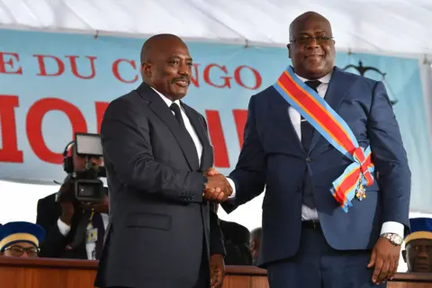 Getty Two men wearing navy suits, white shirts and navy ties shake hands smiling into the camera with the man on the right wearing a sky blue, red and yellow sash