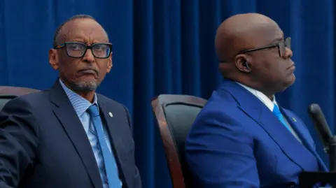 Getty Two men in navy blue suits, white shirts, and blue ties are sitting at a table and looking in opposite directions 