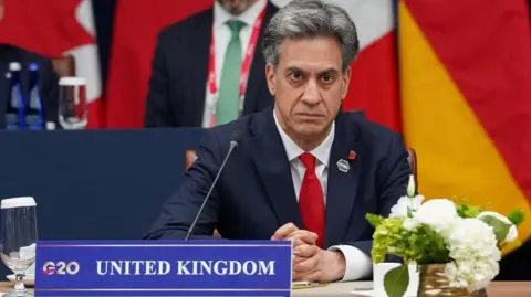 Reuters UK Foreign Secretary Ed Miliband seated at a desk with a sign next to him that reads: G20 United Kingdom. He has his hands clasped and a stern look on his face. 