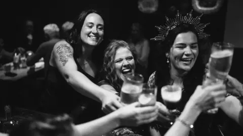 Rob Dight / Epic Love Photography Black-and-white photo of several people gathered around a table, raising pint glasses in a toast. One woman wearing a decorative crown-like headpiece sits near the centre, while others lean in with outstretched arms holding drinks. The indoor setting is softly lit, with blurred figures and tables visible in the background.
