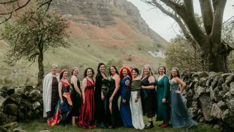 Rob Dight / Epic Love Photography Eleven women wearing colourful dresses stand between two dry stone walls in a rural valley. Trees frame the group on either side, while a steep rocky hillside rises in the background.