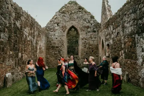Rob Dight / Epic Love Photography A group of women in colourful dresses move through the roofless stone interior of a historic ruin. One person stands near the centre in a green dress and black cloak, while others walk around her, creating a sense of motion. Tall stone walls and a pointed arched window frame the scene, with grass covering the ground inside the building.
