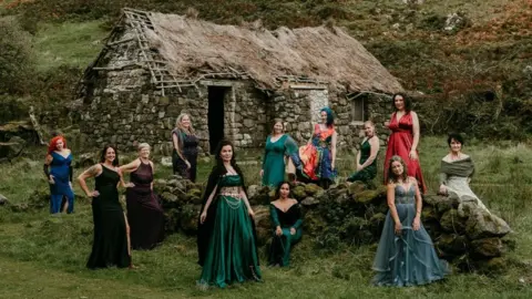Rob Dight / Epic Love Photography A group of 11 women in colourful gowns pose around a low stone wall in front of a traditional stone cottage with a thatched roof. The cottage sits in a grassy rural landscape surrounded by trees and hillside vegetation, while the people are spaced across the scene in various standing and seated positions.
