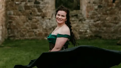 Rob Dight / Epic Love Photography A woman with long brown hair in an off-the-shoulder green dress and dark flowing skirt stands on a grassy lawn in front of a stone building. The skirt billows outward in motion,  stone wall forms the backdrop.