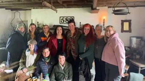 Bridget Devaney A group of 11 women, all wearing colourful jumpers, standing together in a cottage environment smiling widely at the camera.