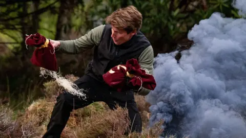 BBC/Studio Lambert/Euan Cherry James Blunt pulling two red money bags out of some grey smoke in some woods
