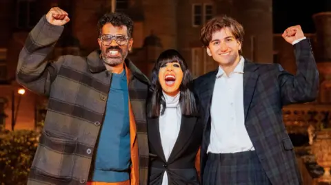 Paul Chappells/ Studio Lambert/BBC Romesh Ranganathan, Claudia Winkleman and Sebastian Croft with their arms around each other and celebrating