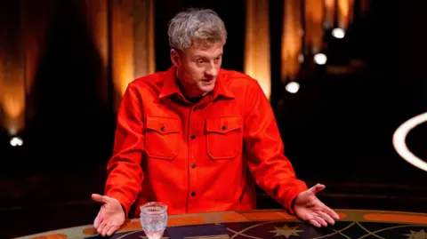 Euan Cherry/Studio Lambert/BBC James Acaster speaking at the round table with both palms outstretched