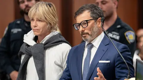 Getty Images Two people in a courtroom, the woman to the left has short cropped blonde hair, a sad expression and is wearing a white hoodie. The man on the right has dark hair, a scruffy beard, and is wearing a blue blazer.
