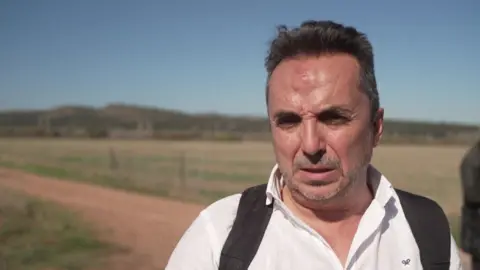 Reuters A man with short hair and a beard - with a bruise on his forehead - speaks to the media; he is wearing a white shirt with the black straps of what appears to be a backpack visible