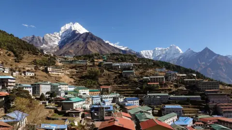 Didier Marti Namche Bazaar's colourful rooftops spread across terraced hillsides in Nepal's Everest region, with the snow-capped peak of Mount Thamserku towering above the town under a clear blue sky.