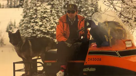 Supplied/Scott Ward An image showing Scott Ward sitting on a snowmobile with a German Shepherd beside him. He is wearing an orange coat with a park warden hat and dark pants. Behind him are snowy trees.