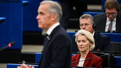 AFP via Getty Images An image showing European Commission President Ursula von der Leyen looking at an out of focus Mark Carney as he delivers a speech to the European Parliament. She has short blonde hair and is wearing a maroon jacket. 