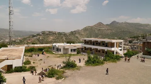 Liam Weir / BBC A school complex made up of low, white buildings positioned around a yard. There are about 20 people dotted around the foreground, and there are mountains in the background. 