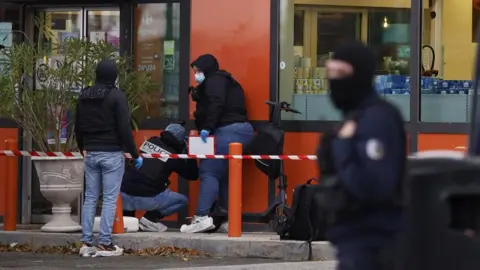 EPA Three police officers crouch down near the wall of a shop. Red and white tape secures the area. Another officer wearing a black balaclava can be seen in the foreground.