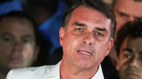 Reuters Brazilian presidential candidate Flavio Bolsonaro of the Liberal Party speaks after losing the presidential runoff election, in Brasilia on 25 October, 2026. He is wearing a white shirt with an open collar. He is gesturing with one hand. Behind him, his supporters look on.