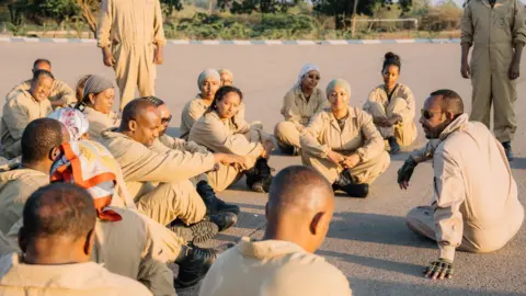 Abiy Ahmed A photo of Abiy Ahmed seated on the ground, while wearing brown military attire. He is speaking to a group of people wearing same brown attire, who are also seated on the ground.