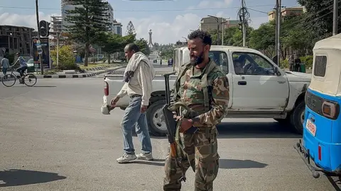 Getty Images A member of the Ethiopian pro-government Tigray Peace Force (TPF) monitors activity on a street while holding a gun. He is in a camo jacket and trousers. A man wearing a white jumper and jeans walks behind him.