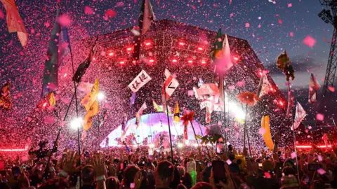 Getty Images The Pyramid stage at Glastonbury festival is pictured lit up with a pink hue at night. There are thousands of pieces of confetti flying through the air as people in the crowd wave their arms and hold up tall flags. 