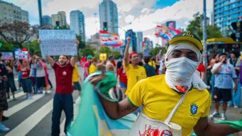 BBC A young man covering his face in a white cloth wearing a yellow Brazil football shirt waving a Brazil flag. In the Background protesters hold up flags and signs.