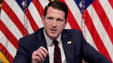 Getty Images A man in a navy-blue suit, white shirt, and blue-and-red striped tie speaks into a microphone. A small rectangular gold pin is attached to his lapel, with two American flags standing behind him.