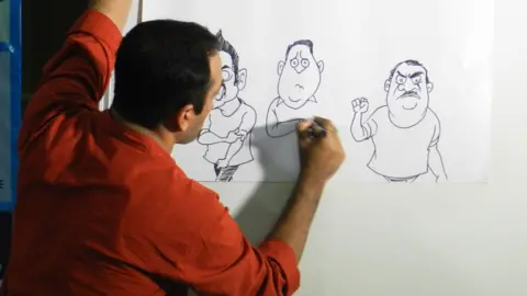 Satish Acharya Satish Acharya wearing a rust shirt draws cartoons on a piece of paper attached to a white board 