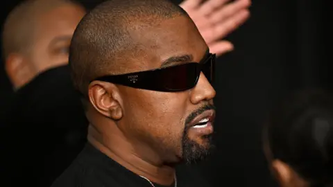 Getty Images Kanye West in 2025 at the Grammy Awards. He has a shaved heard and a black goatee beard. He wears dark sunglasses and a black T-shirt 