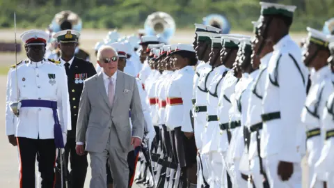 Reuters King Charles, wearing a grey suit and sunglasses, arrives at Lynden Pindling International Airport.