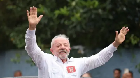 EPA/Shutterstock Luiz Inacio Lula da Silva greets supporters after casting his ballot. He is smiling and waving as he stands through the sunroof of a car. 