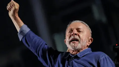 Reuters President Lula extends his right arm, his hand in a fist, as he reacts to his election. He is wearing a blue denim shirt, and has short grey hair and beard.