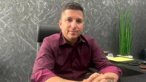BBC Ismael Labrador sits with his hands folded on his desk, wearing a maroon button-down shirt